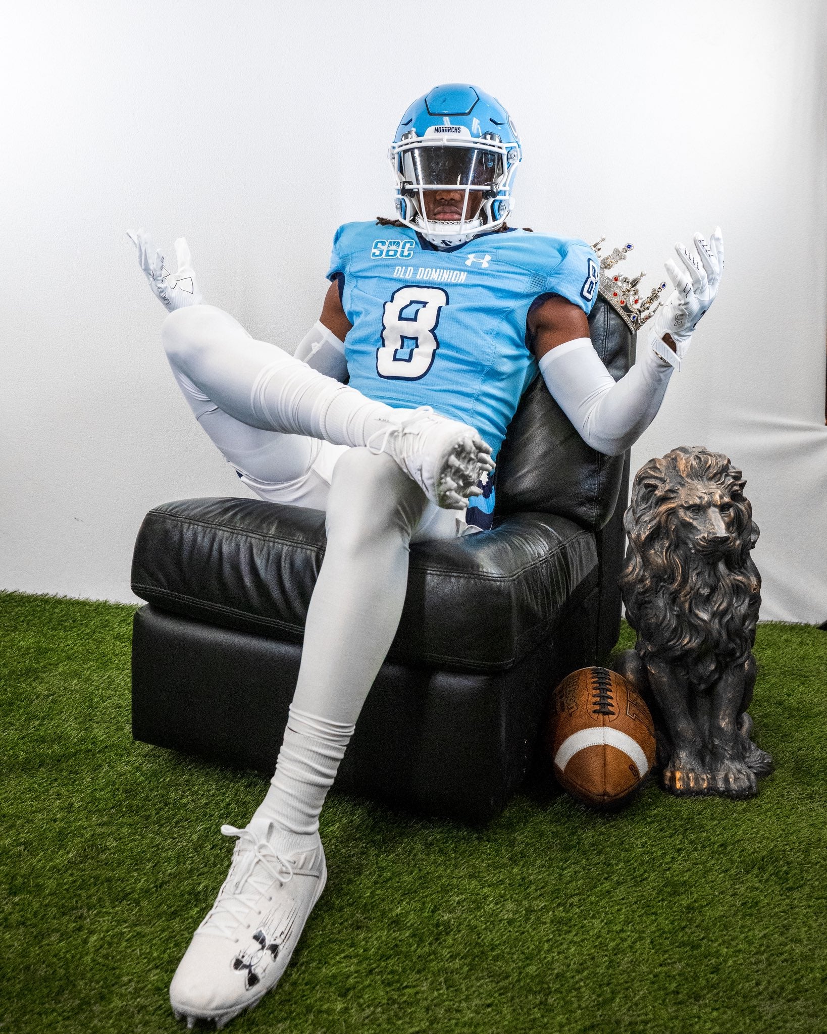 Person in a football uniform sitting on a black leather chair with a football and statue of a dog on a grassy floor.